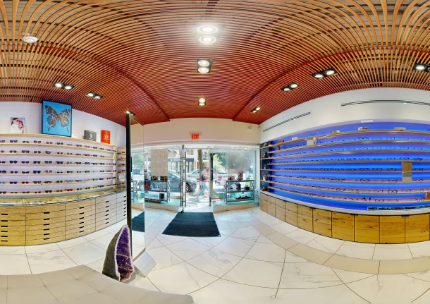 Interior of Envy Eyewear store showing a wide view with wood-slat curved ceiling, backlit display shelves of glasses lining both walls, and the storefront entrance in the background.