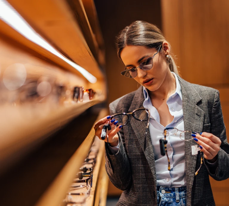 Woman trying on and comparing eyeglass frames at a display wall in an optical store.