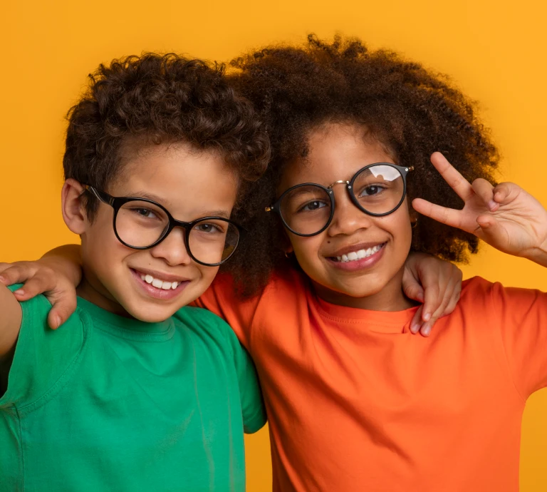 Two smiling children wearing round eyeglasses, standing arm-in-arm against a bright yellow background, one making a peace sign.