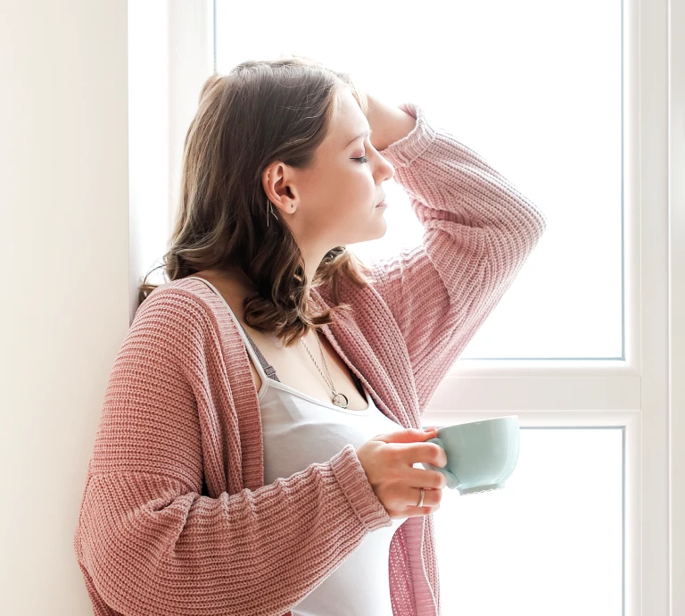 Woman in a pink cardigan leaning against a wall by a bright window, eyes closed, holding a mug of coffee or tea.