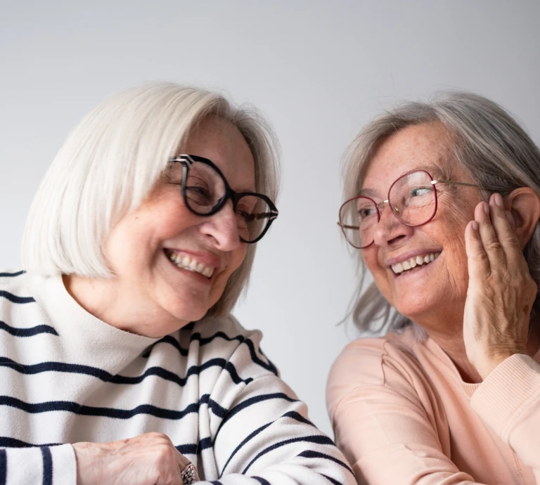 Two smiling older women wearing eyeglasses, one in round black frames and the other in round wire frames, laughing together against a neutral background.
