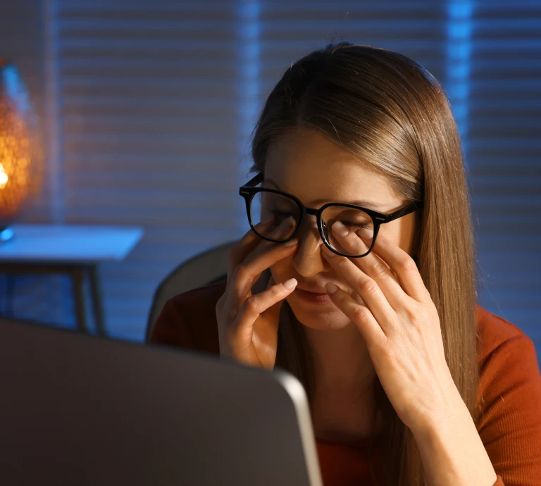 Woman wearing glasses rubbing her tired eyes while sitting in front of a laptop screen, illustrating digital eye strain.