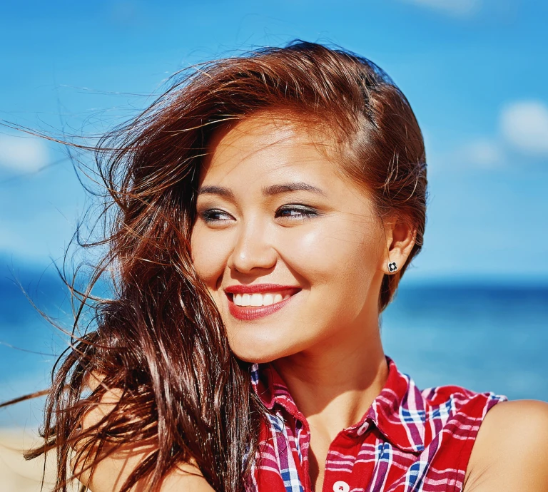 Close-up portrait of a smiling woman with windswept hair, standing outdoors against a blue sky and ocean backdrop.