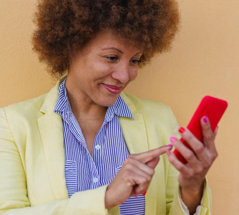 Smiling woman with curly hair looking at her phone, wearing a striped shirt and yellow blazer against a warm-toned background.