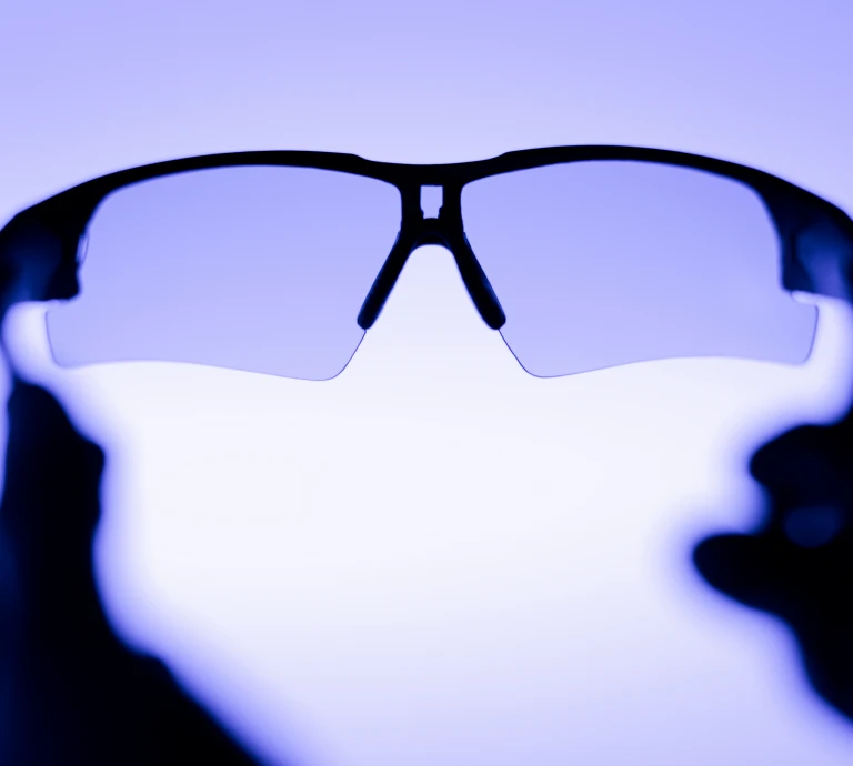 Close-up of sport-style sunglasses with wraparound lenses, held up against a blue-toned light with hands visible in silhouette on either side.